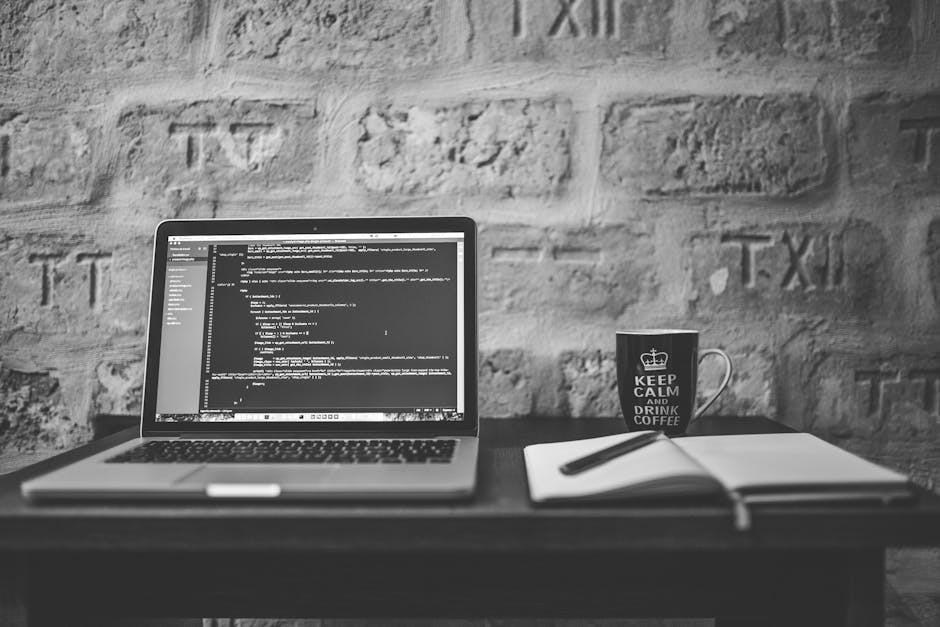 A workspace featuring a laptop displaying lines of code on a black screen, placed on a dark desk against a textured brick wall. Next to the laptop, there is an open notebook with a black pen resting o