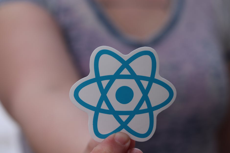Close-up of a person's hand holding a sticker featuring a blue atomic model symbol in front of their chest, with the background showing a blurred figure wearing a purple top. The setting appears to be